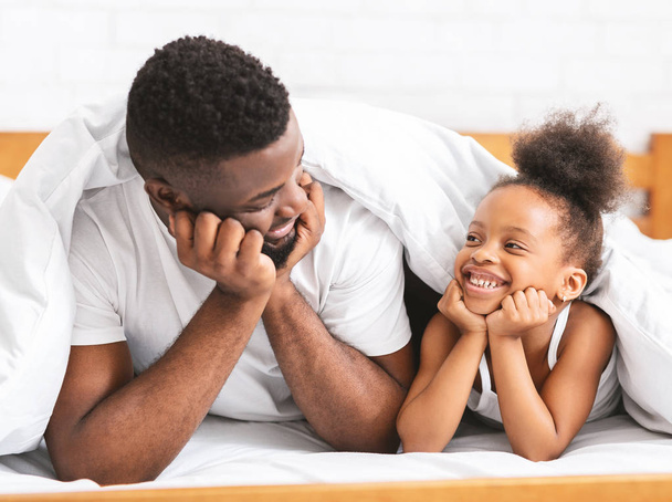 süße afrikanische Vater und Tochter liegen auf dem Bett und lächeln einander zu - Foto, Bild