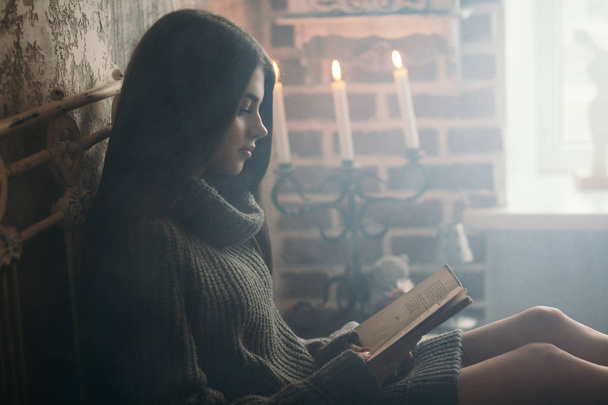 Beautiful young woman in knitted sweater sitting on bed and reading book near window at home. Winter atmosphere - 写真・画像