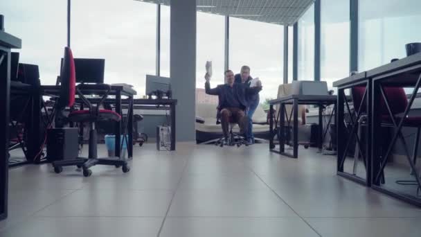 Funny crazy businesspeople young men are having fun with rolling chair, one businessman is pushing armchair other is moving forward in office hall. Youth and joy in succesfull business concept - Footage, Video