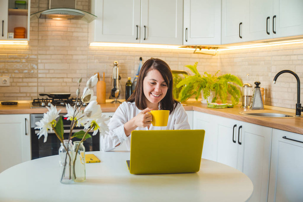 жінка п'є чай з жовтої чашки, що працює на ноутбуці. кухня на фоні
 - Фото, зображення