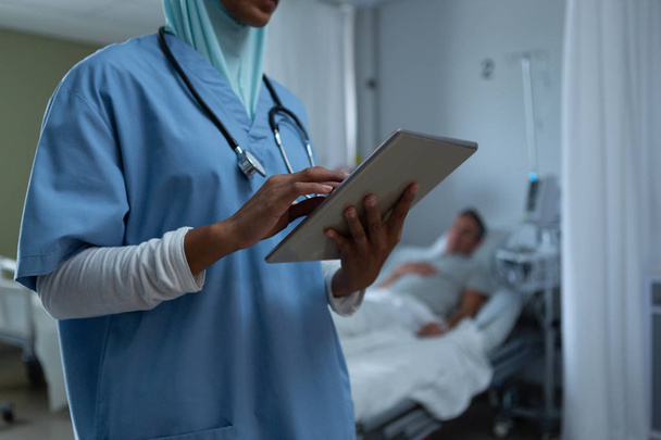 白人男性の患者が病院でバックグラウンドで眠っている間、病棟でデジタルタブレットを使用してヒジャーブでかなり混合人種の女性医師の中間セクション - 写真・画像