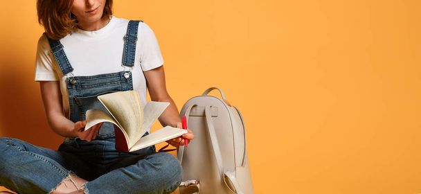 Portrait of a pensive girl in a white T-shirt and denim overalls, sitting on the floor with a red notebook and a pencil on a yellow background - Foto, Imagem