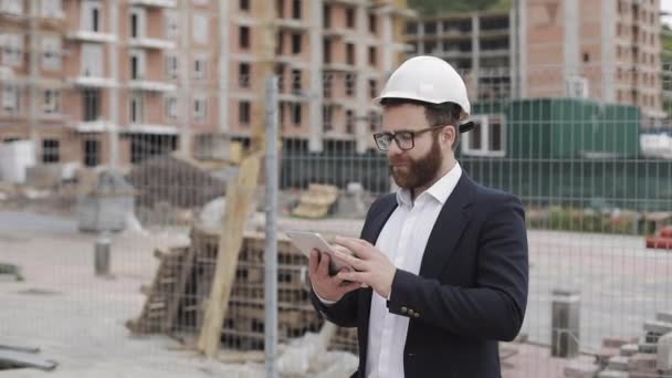 Portrait of young businessman with tablet on construction site smiling looking to camera wearing a safety helmet and suit. Engineer, architect, builder, businessman. - Filmati, video