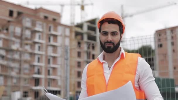 Portrait of construction worker in orange helmet looking at the camera. The builder with construction project stands on the construction site background. Slow motion. - Πλάνα, βίντεο