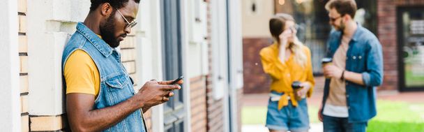 外のスマートフォンを使用してサングラスでハンサムなアフリカ系アメリカ人男性のパノラマショット  - 写真・画像