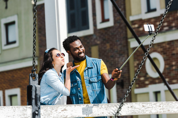 attrayant femme envoi air baiser près beau afro-américain homme prendre selfie tout en tenant selfie bâton
  - Photo, image