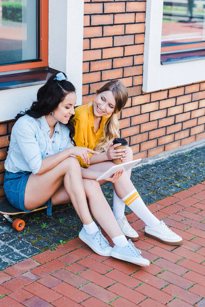 fröhliche Frau zeigt mit dem Finger auf digitales Tablet, während sie mit Freund auf dem Penny-Board sitzt  - Foto, Bild