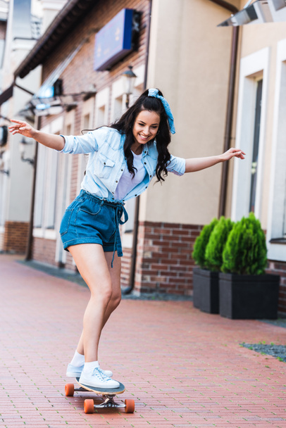 happy young woman with outstretched hands riding penny board near building  - Foto, immagini