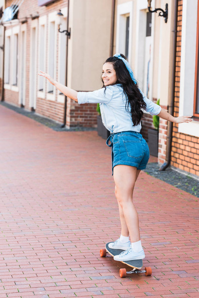 cheerful young woman with outstretched hands riding penny board near building  - Foto, Bild