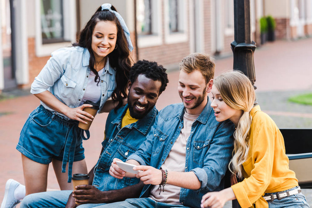 amici multiculturali felici guardando smartphone mentre seduti sulla panchina
  - Foto, immagini