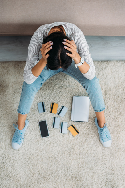 high angle view of upset woman sitting on floor with credit cards, notebook and smartphone  - Photo, Image