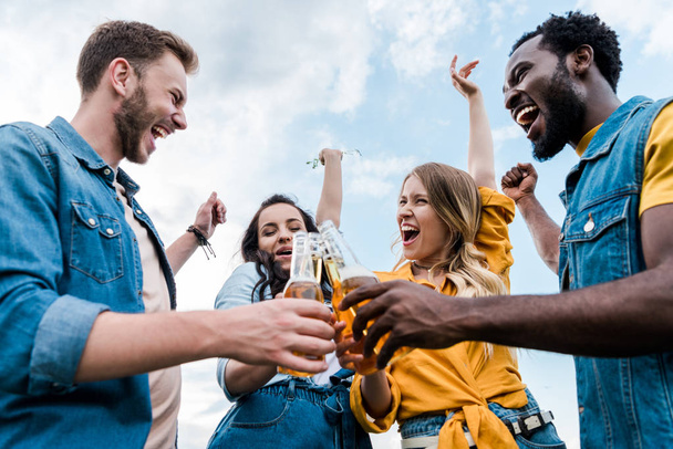 low angle view of happy multicultural men and women clinking bottles with beer  - Fotografie, Obrázek