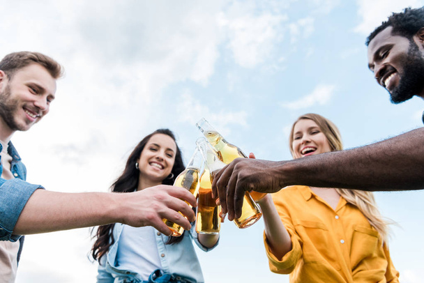 low angle view of cheerful multicultural men and women clinking bottles with beer  - Foto, Bild