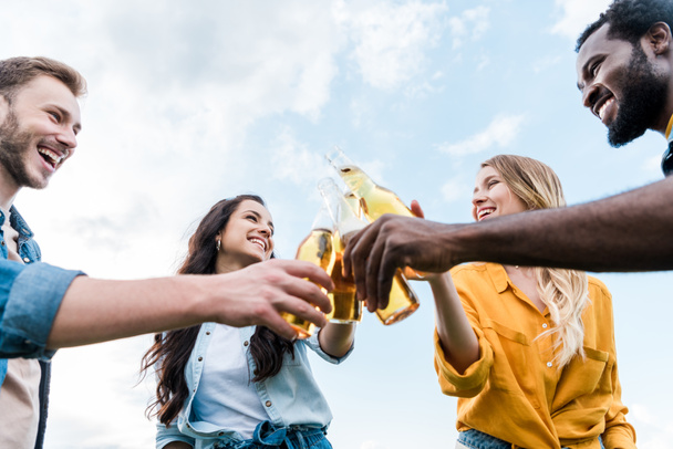 low angle view of cheerful multicultural women and men clinking bottles with beer  - Fotoğraf, Görsel
