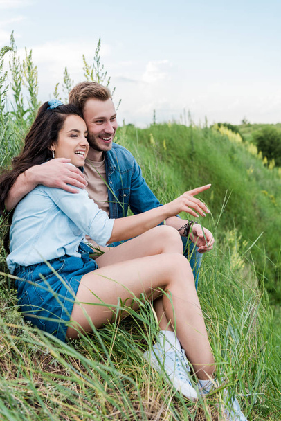 緑の草の上にハンサムな男性と座って、指で指を指している陽気な若い女性の選択的な焦点  - 写真・画像