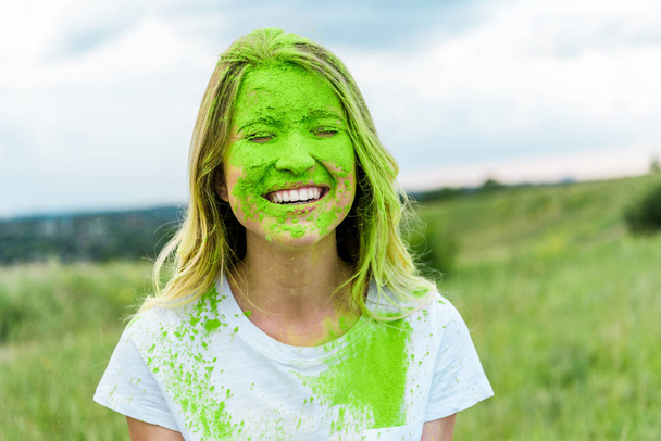 vidám nő csukott szemmel és zöld Holi festéket arcán mosolygós szabadban  - Fotó, kép