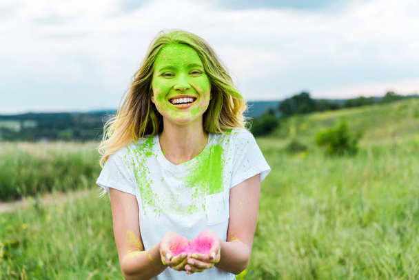cheerful woman with green holi paint on face standing with cupped hands and smiling outdoors  - Photo, Image