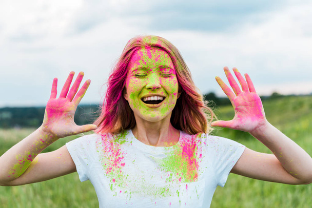 cheerful woman with closed eyes and pink holi paint on hands gesturing and smiling outdoors  - Photo, image