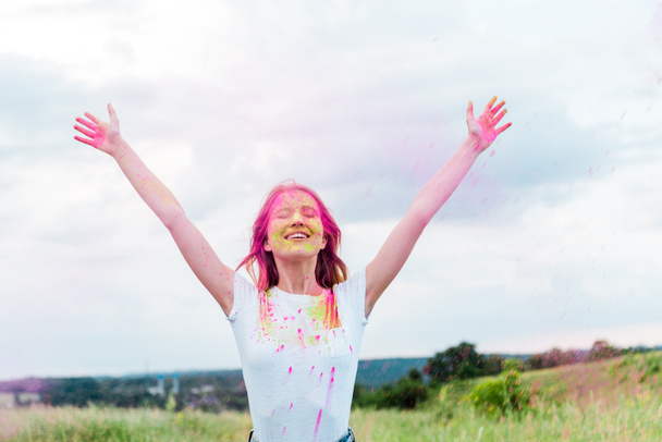 happy woman with closed eyes and pink holi paint on outstretched hands smiling outdoors  - Photo, Image