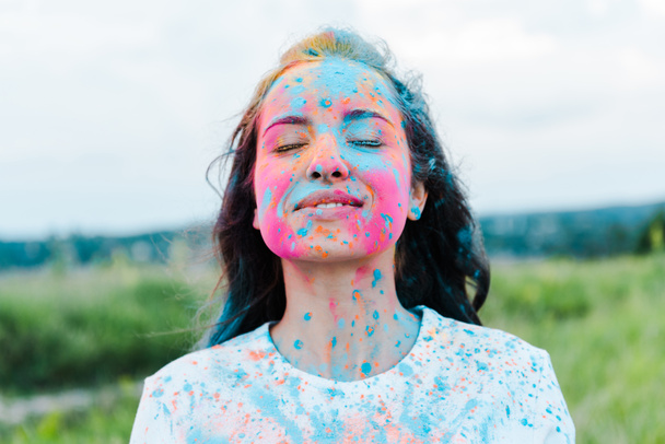 positive woman with closed eyes and colorful holi paints on face  - 写真・画像