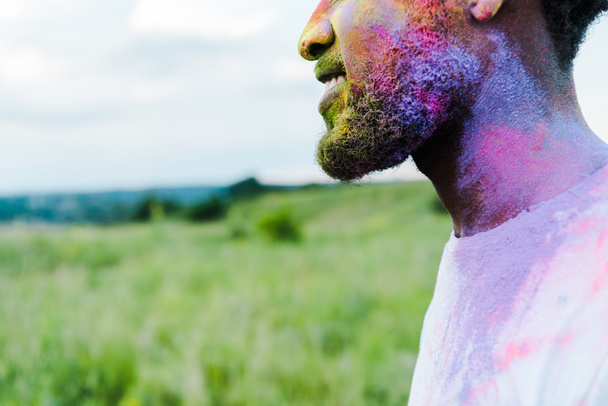 обрізаний вид веселого афроамериканського чоловіка з барвистими фарбами холі на обличчі
  - Фото, зображення