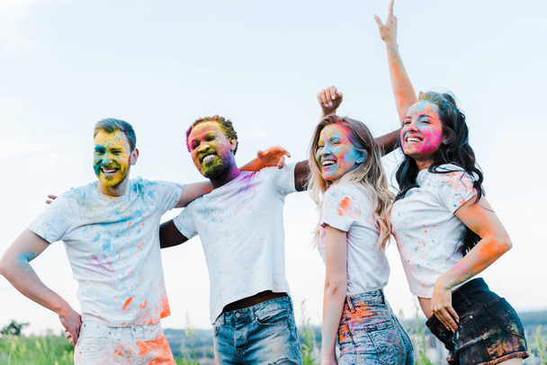 happy man with hand on hip near multicultural friends with holi paints on faces  - Photo, Image