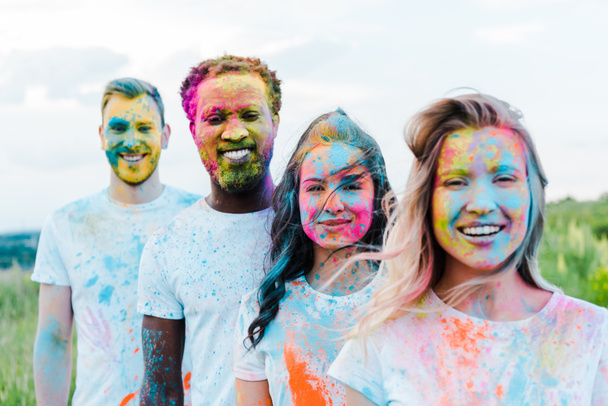 selective focus of woman smiling near multicultural friends with holi paints on faces  - 写真・画像