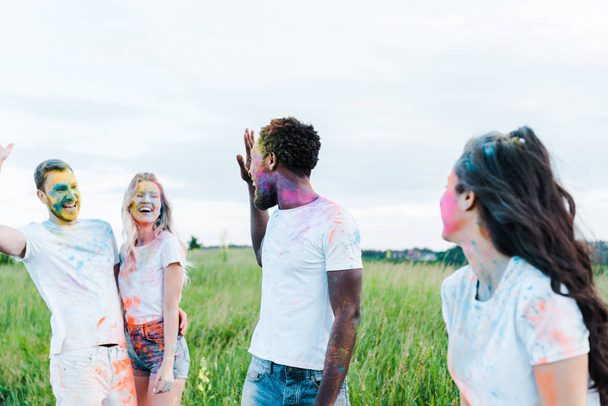 selective focus of multicultural friends with holi paints on faces waving hands  - Foto, Imagem