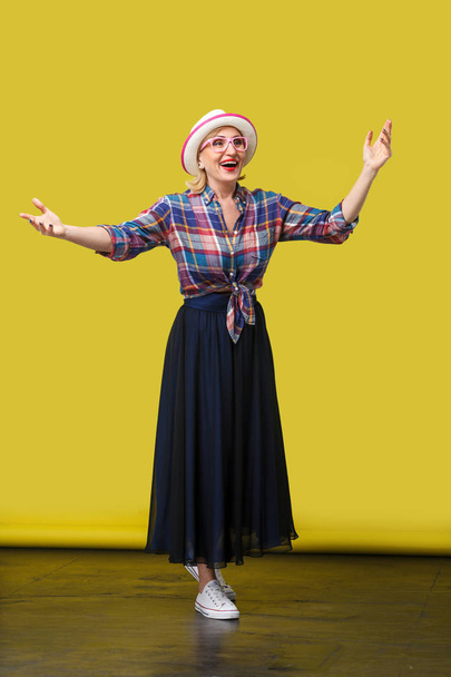 happy modern stylish mature woman in casual style with hat standing with raised arms and surprised face on yellow background - Fotografie, Obrázek