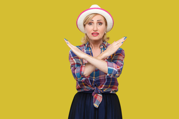 ernstige moderne stijlvolle volwassen vrouw in casual stijl met hoed toont X teken en kijken naar camera op gele achtergrond - Foto, afbeelding