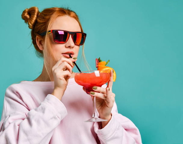 Stylish blonde woman in modern red sunglasses drinks through straw a strawberry margarita cocktail - Fotó, kép