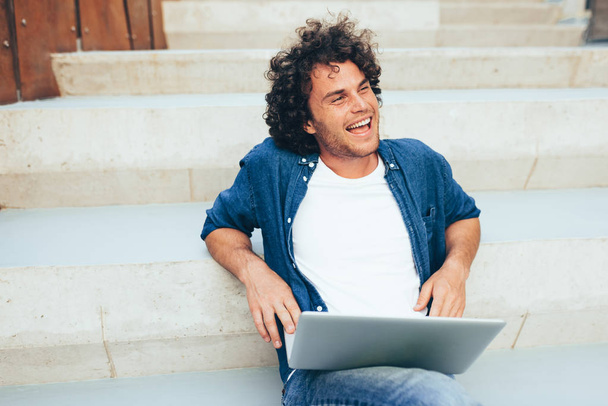 Outdoors shot of cheerful male with curly hair using laptop for chatting online with friends, connected to free wireless on the city street. Handsome man texting something on his laptop computer. - Zdjęcie, obraz