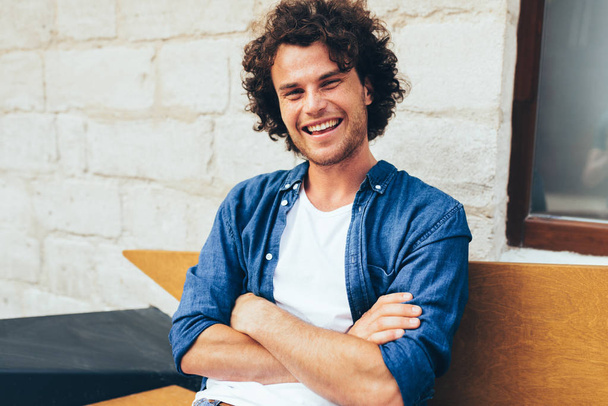 Handsome young man with curly hair, looking to the camera, smiling broadly, sitting on the bench in the street. Happy male student relaxing with keeps arms crossed outdoors. People, lifestyle concept - Фото, изображение
