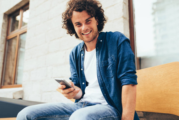 Smiling young man stitting outdoors texting on mobile phone. Happy male with curly hair using smart phone application for searching cafes near location in downtown in the city. Lifestyle and people - Photo, Image