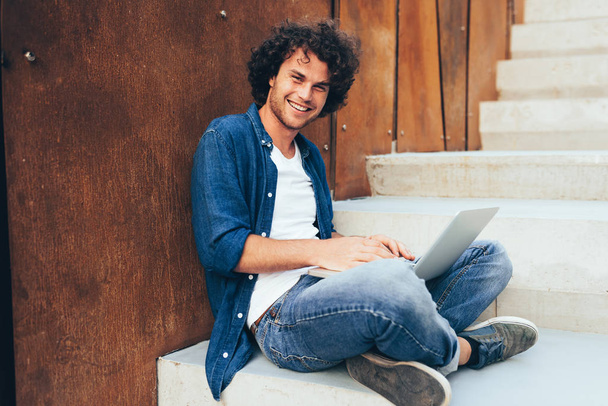 Happy man with curly hair using laptop for chatting online with friends, browsing on Internet on the city. Handsome male smiling broadly sitting on the concrete stairs, learning on his laptop computer - Valokuva, kuva