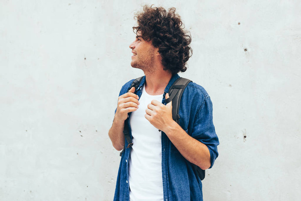 Horizontal side view portrait of Caucasian young man with curly hair, smiling, with backpack on the back, standing at building concrete background on the city street. People and lifestyle concept. - Fotografie, Obrázek