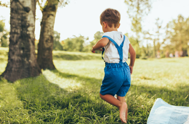 Rear view image of happy little boy wearing white t-shirt and blue shorts  playing at nature background. Cheerful child running on the green grass in the park. Kid having fun outdoors. Childhood. - Φωτογραφία, εικόνα
