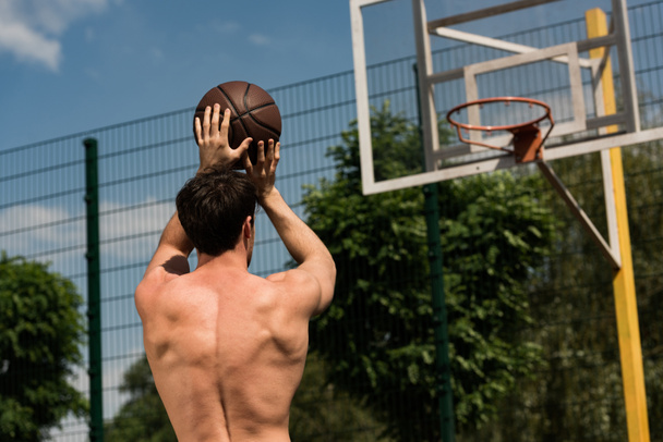 back view of shirtless basketball player throwing ball in basket at basketball court - Foto, afbeelding