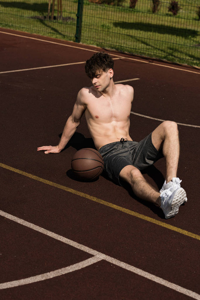sexy shirtless basketball player with ball sitting at basketball court - Zdjęcie, obraz
