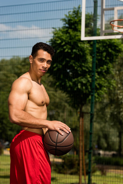 shirtless mixed race basketball player with ball looking at camera at basketball court - Foto, Bild
