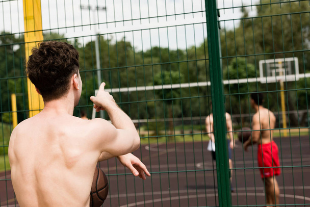 Вид сзади на баскетболиста с мячом, смотрящего на друзей и указывающего пальцем на баскетбольную площадку
 - Фото, изображение