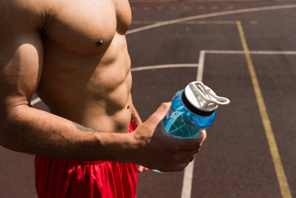partial view of shirtless muscular sportsman holding sport bottle at basketball court - Foto, imagen