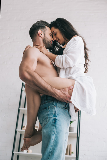 przystojny mężczyzna w dżinsy gospodarstwo i całowanie z seksowny Kobieta w koszula  - Zdjęcie, obraz
