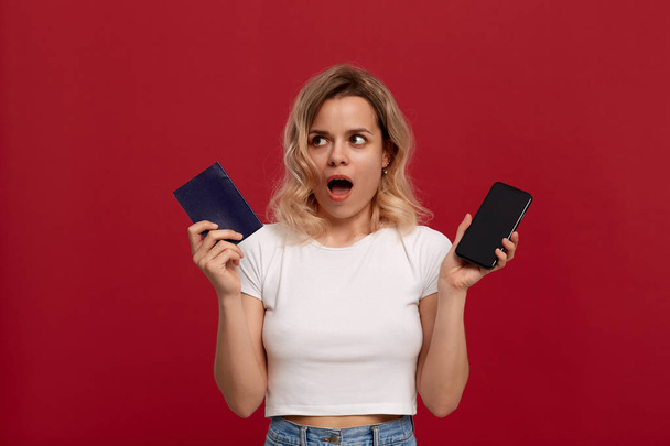 Porträt eines Mädchens mit blonden Locken in einem weißen T-Shirt auf rotem Hintergrund. Überraschtes Model blickt in die Kamera und hält Reisepass mit Handy in der Hand. - Foto, Bild