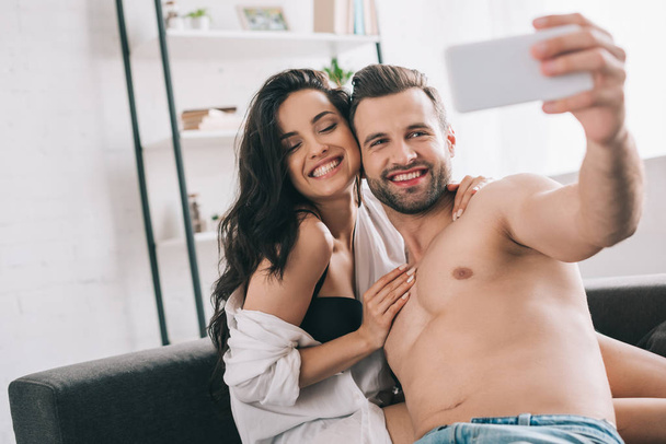 όμορφος άντρας και σέξι γυναίκα σε πουκάμισο και σουτιέν χαμογελώντας και παίρνοντας selfie  - Φωτογραφία, εικόνα