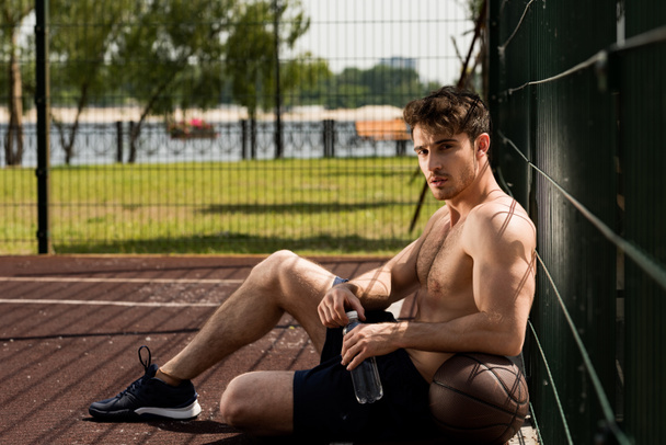 Sexy muskularny sportowiec z piłką i butelką wody siedzącej na boiska do koszykówki - Zdjęcie, obraz