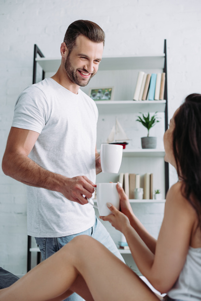 handsome and smiling man giving cup with tea to brunette woman  - Foto, imagen
