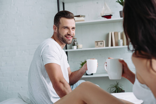handsome and smiling man and brunette woman looking at each other and holding cups - Foto, imagen