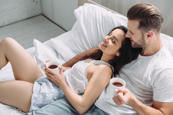 aantrekkelijke en brunette vrouw en man houden kopjes, liggend op bed en knuffelen  - Foto, afbeelding