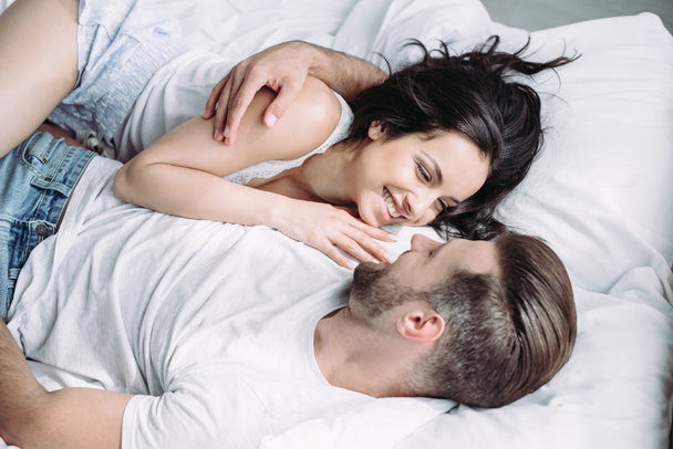attractive and brunette woman and man smiling, lying on bed and hugging  - Zdjęcie, obraz